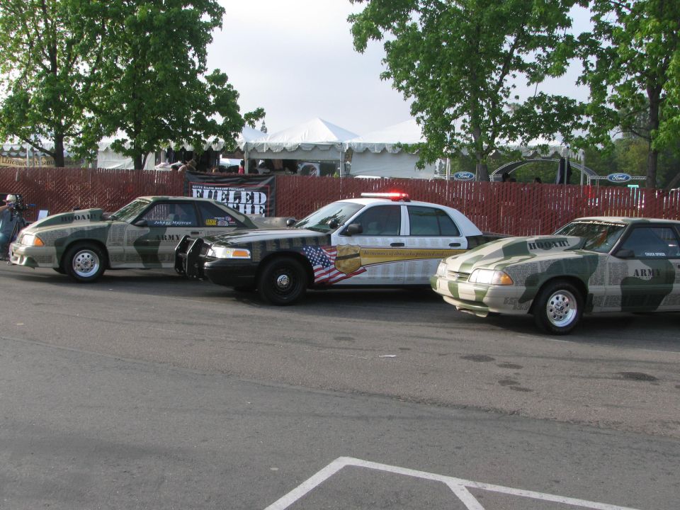 Three cars, two Mustangs and a police car, are parked side-by-side. The Mustangs have camouflage paint and "ARMY" text. The police car has lights on