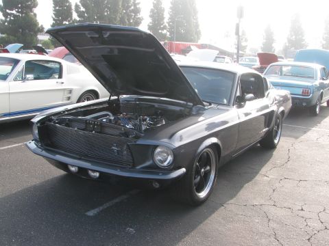 1996 Ford C-350 (Centurion Conversions) - A dark gray 1967 Ford Mustang coupe is parked with its hood open, revealing the engine bay. Other classic vehicles are present in the backg…