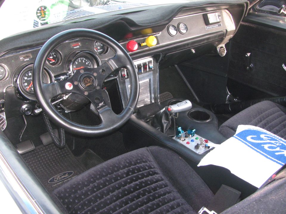 Interior view of a car with a GT steering wheel, custom shifter, and racing gauges. A Ford towel is on the passenger seat
