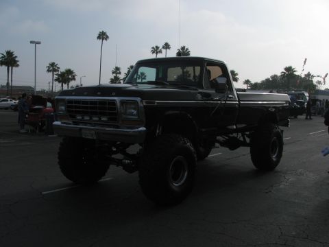 1996 Ford C-350 (Centurion Conversions) - A heavily modified black Ford pickup truck with oversized tires is parked in a lot with palm trees in the background.