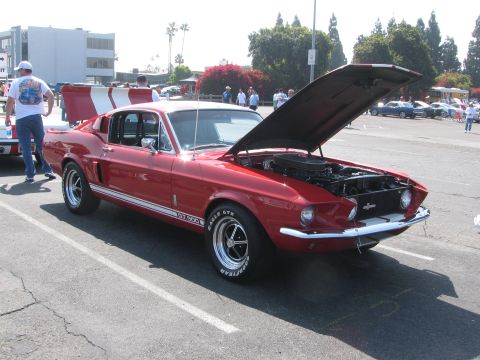 1996 Ford C-350 (Centurion Conversions) - A red 1967 Shelby GT500 is parked with its hood open, displaying its engine. Another car is partially visible to the left.