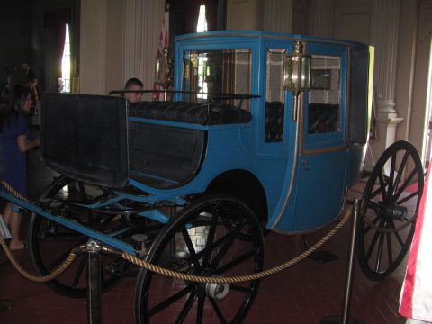 1996 Ford C-350 (Centurion Conversions) - A blue carriage with black wheels and seating is displayed indoors, cordoned off with rope.