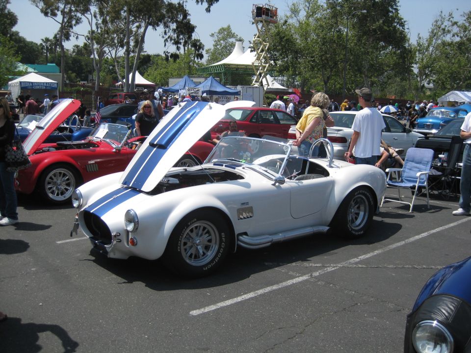 White convertible sports car with blue stripes on hood and trunk, parked at an outdoor event with other cars