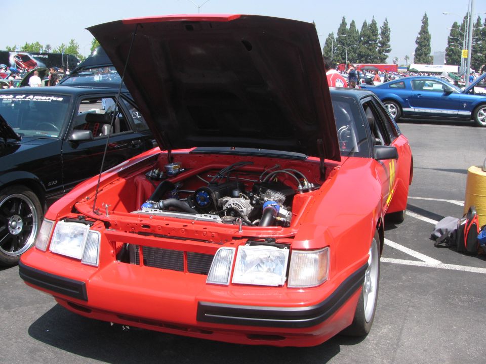 Red car with hood open, engine visible. Black car to the left, blue car in background. Parking lot setting