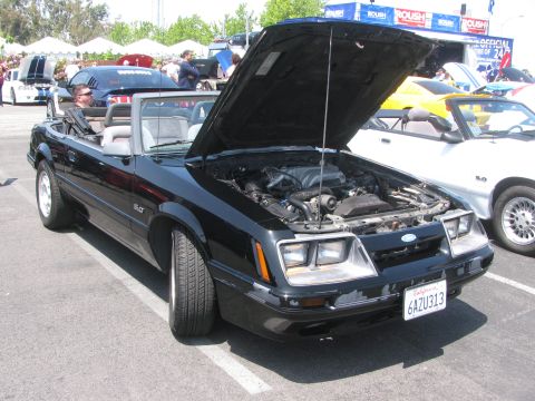 1996 Ford C-350 (Centurion Conversions) - A black convertible Mustang is parked with its hood up, revealing the engine bay. Another white Mustang is visible to the right.