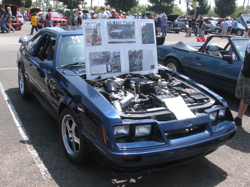 Blue Ford Mustang, hood open showing engine, display board in front of windshield. Other cars and people in background