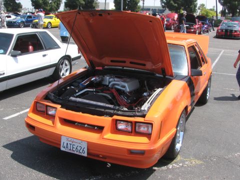 1996 Ford C-350 (Centurion Conversions) - An orange Fox body Mustang is displayed with its hood open, revealing the engine bay, at an outdoor car event.