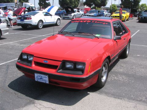 1996 Ford C-350 (Centurion Conversions) - A red Ford Mustang hatchback is parked in a lot with other cars, displaying a "Wicked Stangs" banner.