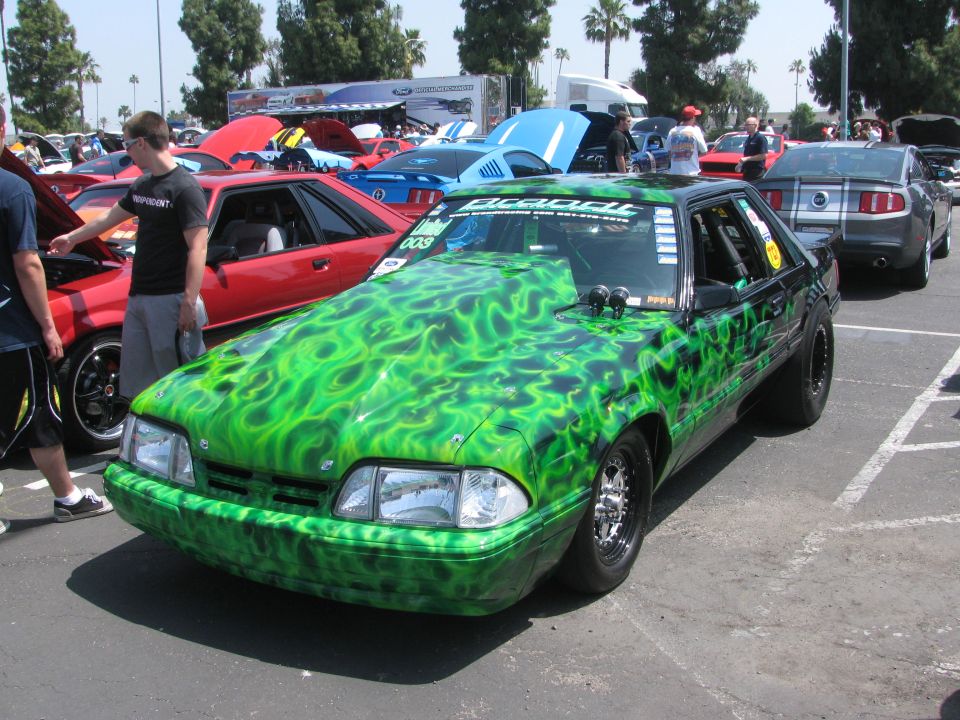 Customized Ford Mustang with green flame paint, drag racing slicks, and hood scoop. Other cars are visible in the background