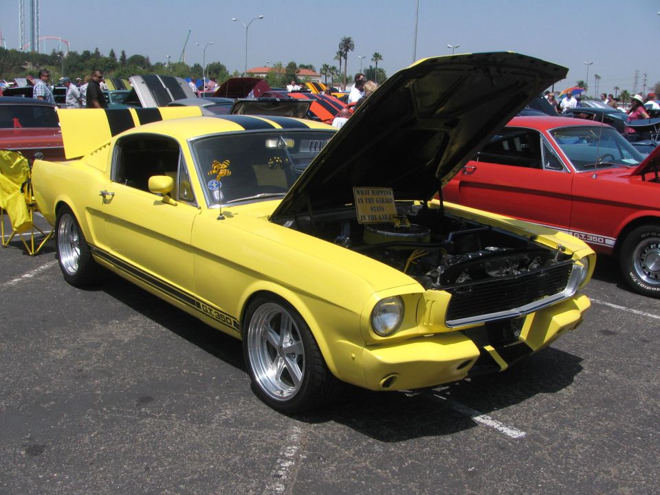 Yellow 1966 Ford Mustang GT350 with black racing stripes, hood open. Another red classic car is parked next to it