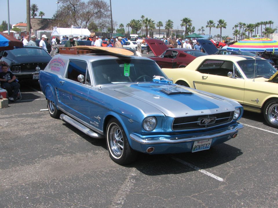 Blue and silver 1965 Ford Mustang "SportsRoof" wagon conversion with surfboard on roof, parked at car show. License plate reads PONYEX2