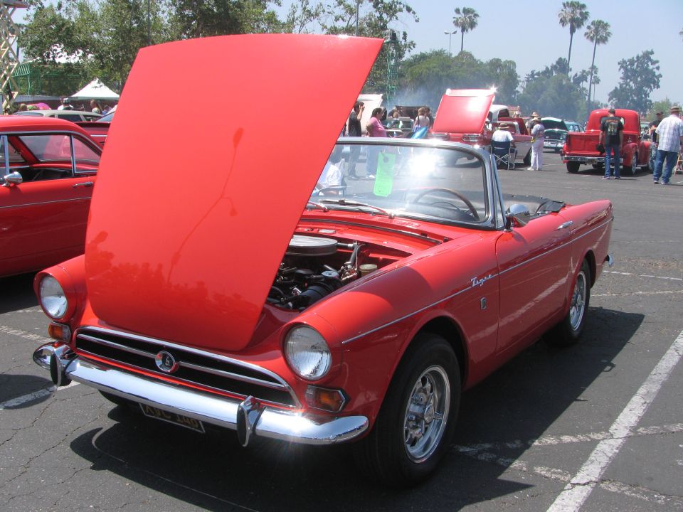 Red convertible sports car with hood open, showing engine. Another red car is parked to the left. Several people and other vehicles are in the background