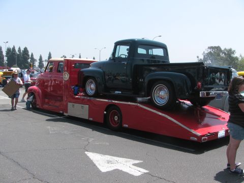 1996 Ford C-350 (Centurion Conversions) - A dark green 1956 Ford F-100 pickup truck is transported on a red car carrier, with another vehicle visible in the background.