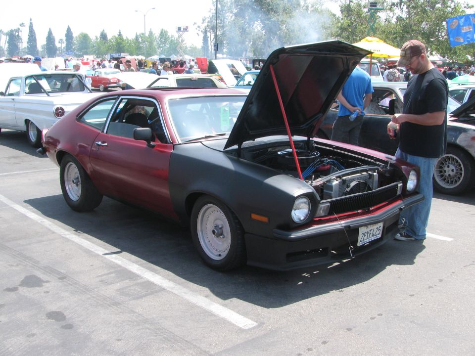 Red and black Ford Pinto coupe with hood open, showing engine. White wheels, custom bodywork