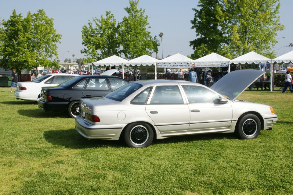 Three Ford Taurus sedans parked on grass, one with hood open. The closest car is silver, the middle is black, and the furthest is white