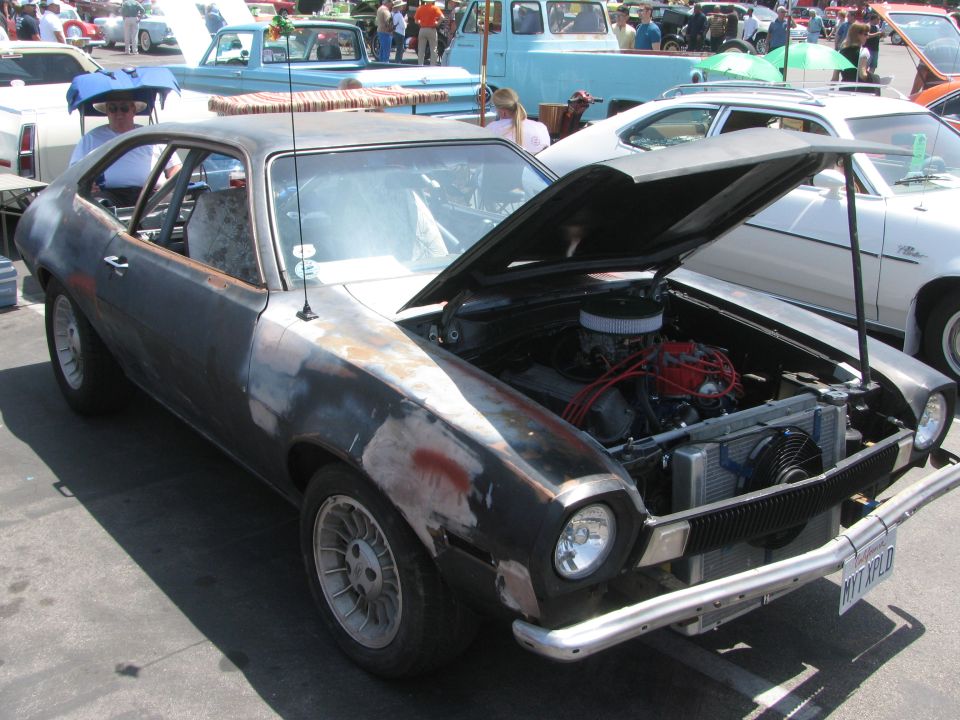 Old Ford Pinto with hood open, showing engine. Body is rusty and has primer patches. Other cars and people in background