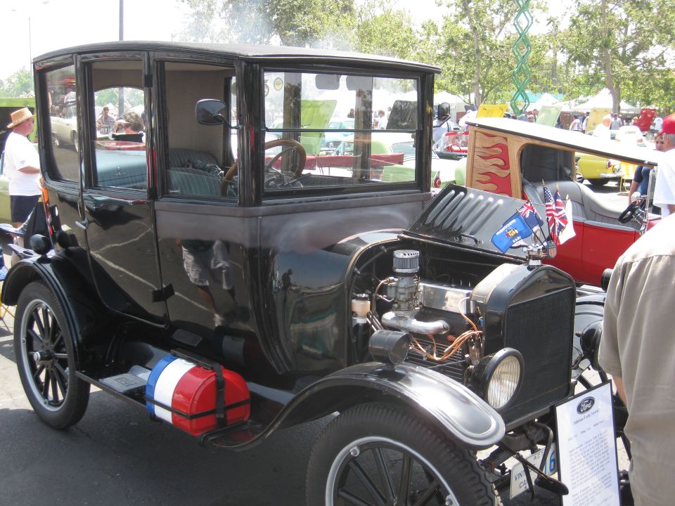 Black antique car with open hood, flags on radiator cap, and a red, white, and blue fuel tank