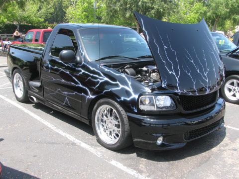 1996 Ford C-350 (Centurion Conversions) - This custom black pickup truck features striking lightning bolt graphics and its hood is open, revealing the engine.