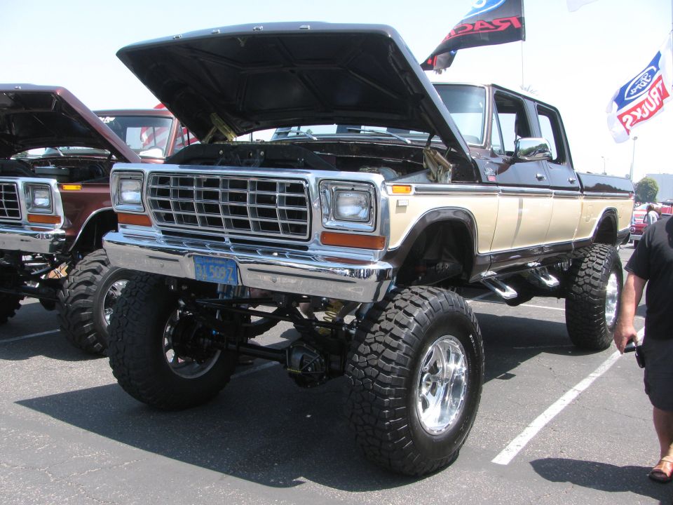 Lifted Ford pickup truck with hood open, another similar truck next to it. Ford flags in background
