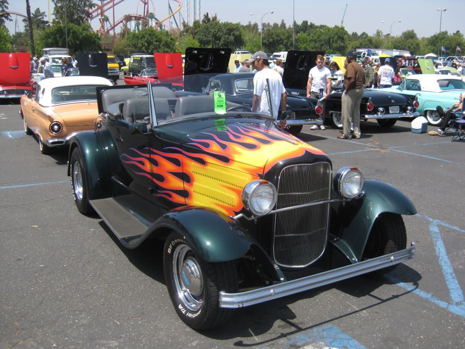 Green hot rod with flames painted on the hood, parked in a lot with other cars and people