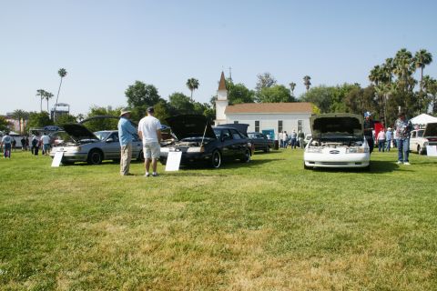 1992 Ford Taurus - A white 1992 Ford Taurus is displayed with its hood open at an outdoor car show, with other vehicles and attendees in the background.