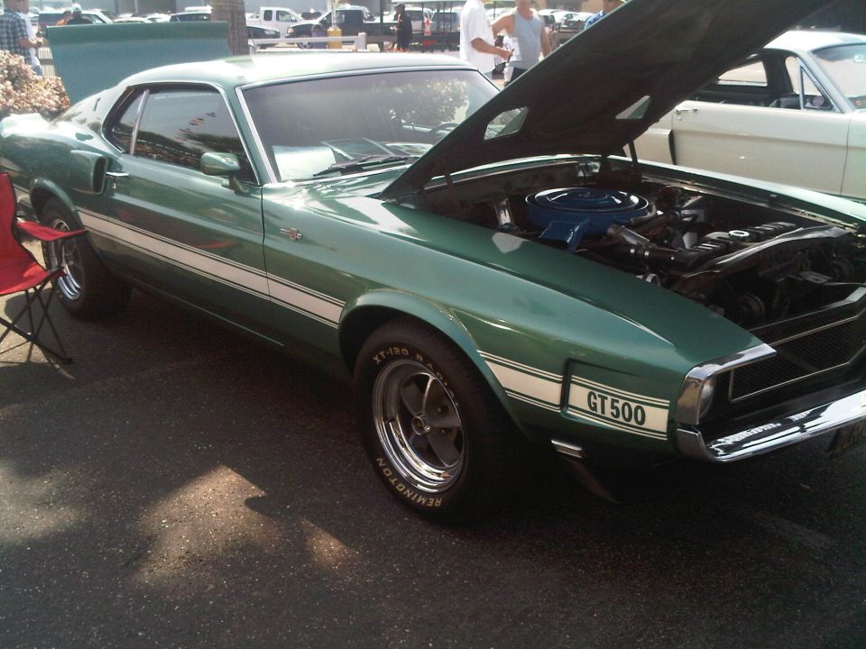 Green 1969 Ford Mustang GT500 with white racing stripes, hood open, showing engine, parked outdoors