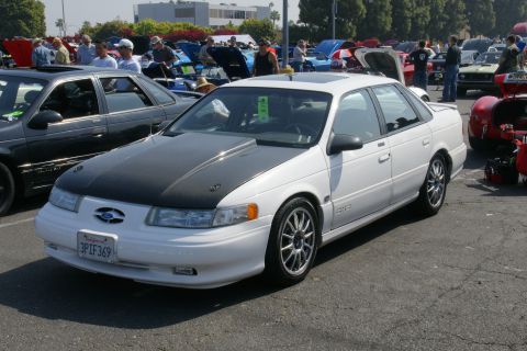 1992 Ford Taurus - A white 1992 Ford Taurus sedan with a black hood and racing stripes is displayed at a car show.