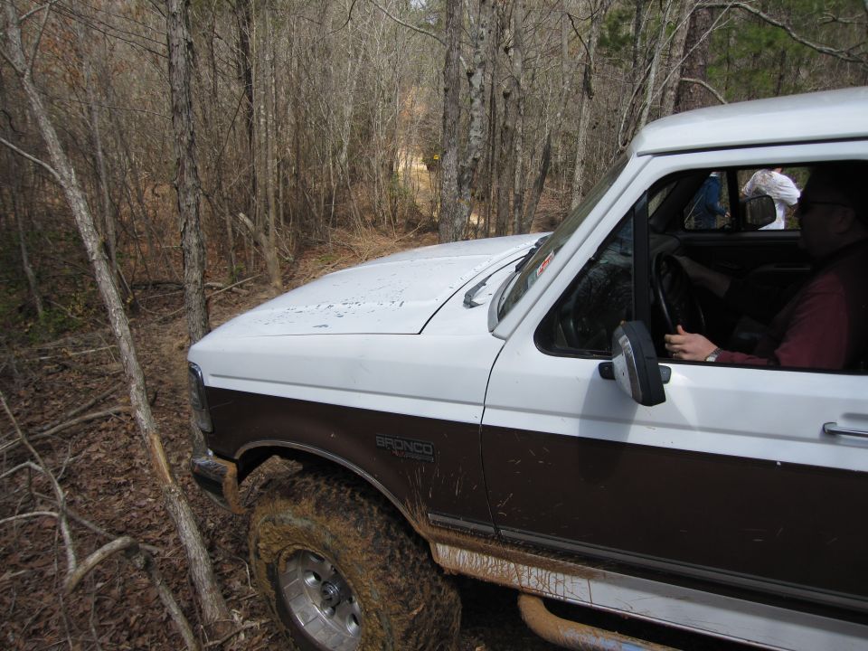 1993 Ford Bronco driving on a muddy, wooded trail. "BRONCO" badge visible on fender