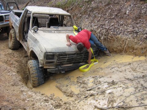 1993 Ford Bronco - A man struggles to free a stuck 1980s Ford Bronco from a muddy trench, with another vehicle visible behind it.