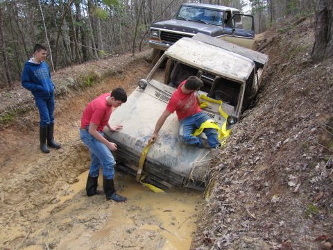 1993 Ford Bronco - Two people attempt to free a white Ford Bronco from a muddy ditch, while a brown Ford pickup truck is positioned to pull it out.