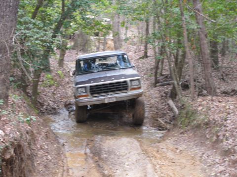 1993 Ford Bronco - This lifted Ford truck navigates a water obstacle on a dirt trail surrounded by trees.