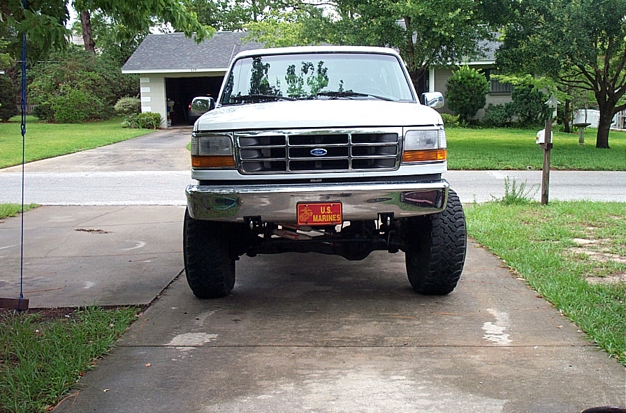 Front view of a white lifted Ford truck on a driveway with chrome bumper and US Marines plate