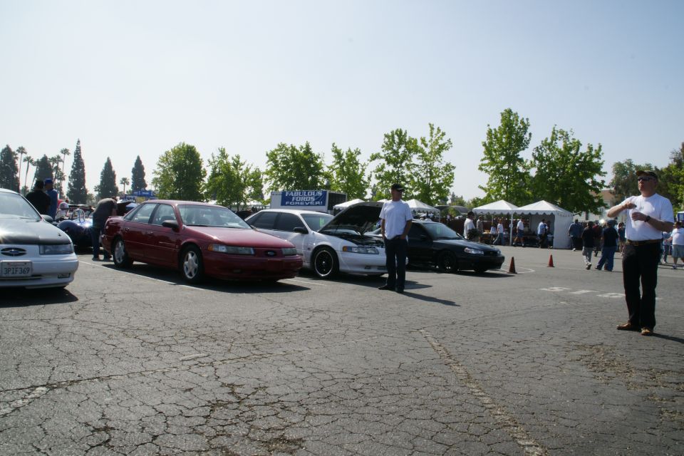 Several Ford Taurus cars parked in a lot, some with hoods up. People are milling around tents in the background