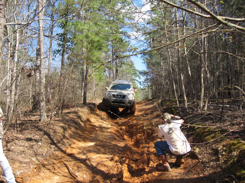 Nissan Xterra climbing a muddy hill in a wooded area. Person crouching to take photo