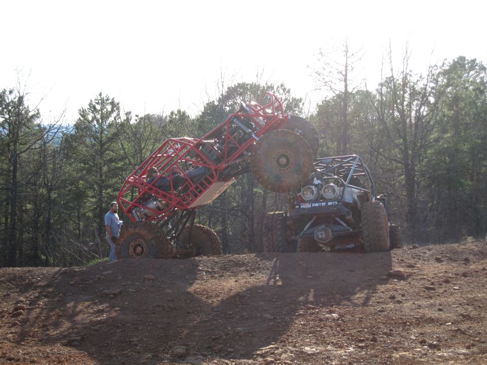 Two custom off-road buggies, one red and one black, are climbing a steep dirt incline with trees in the background