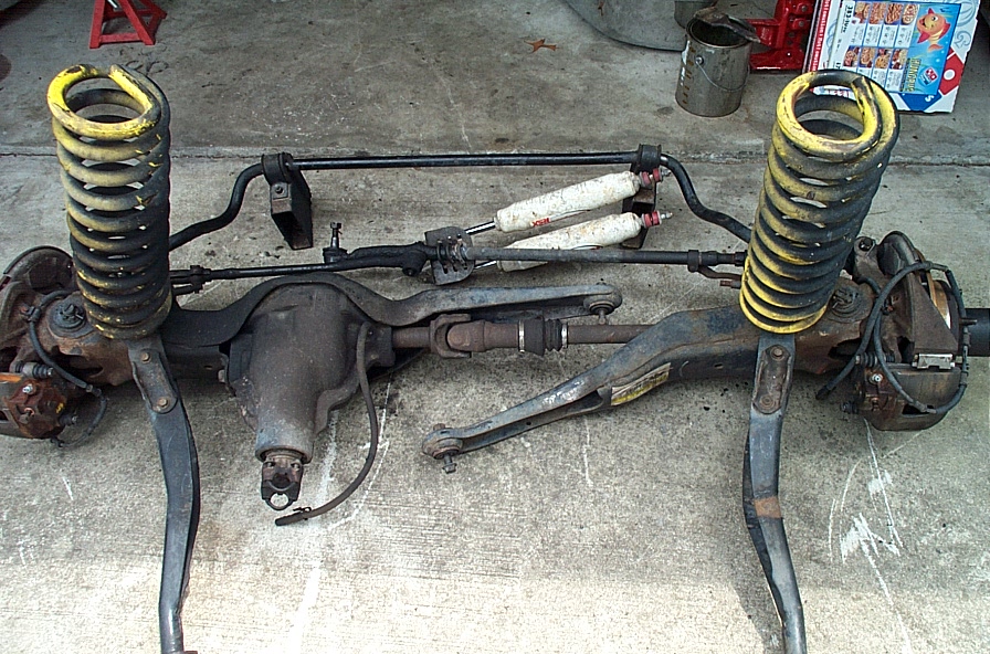 Removed front axle assembly with yellow coil springs, sway bar, and shocks laid on concrete driveway