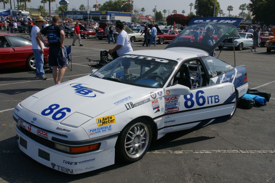 White and blue Ford Probe race car with number 86, hood and trunk open, parked in a lot with other cars and people