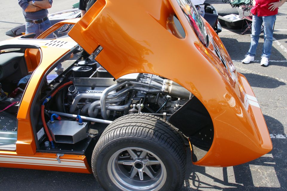 Orange sports car with hood open, showing V8 engine and exhaust headers. White stripes on front fender