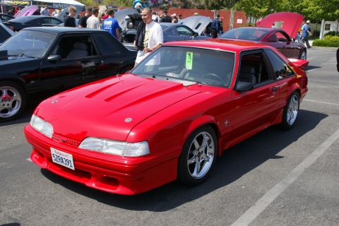 1992 Ford Taurus - A red coupe with a hood scoop and chrome wheels is parked at an outdoor car show.
