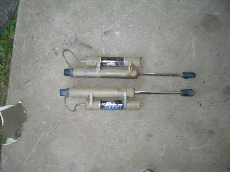 Two Fox shock absorbers with blue ends and silver shafts, lying on concrete