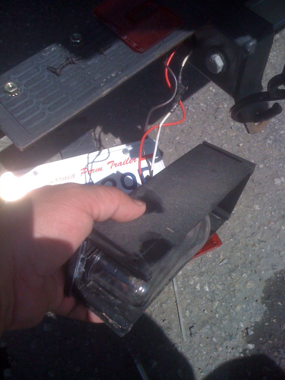 Hand holding a removed trailer tail light housing with spliced red and white wires at the rear crossmember