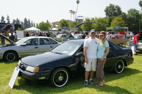 1992 Ford Taurus - A couple poses with a black 1992 Ford Taurus at a car show, with other vehicles and tents in the background.