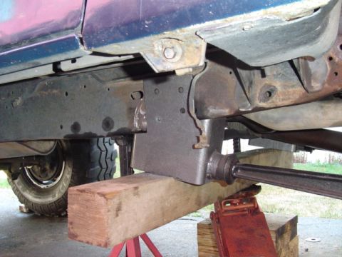 1993 Ford Bronco - Underbody view of a purple/maroon truck body supported on a red jack stand and wooden blocks, showing a bare steel skid plate or crossmembe…