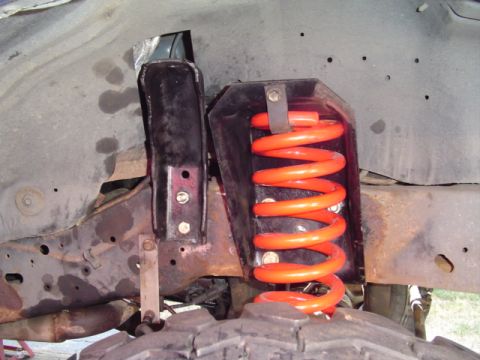 1993 Ford Bronco - Wheel-well view of a bright orange coil spring seated in a fabricated black steel spring bucket bolted to the frame rail, with a knobby off…
