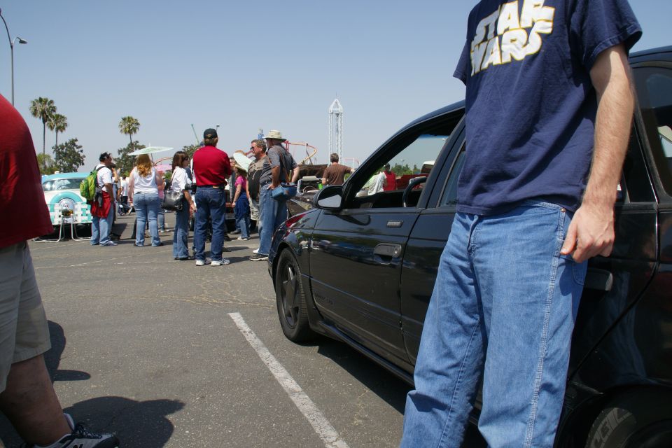 guy in Star Wars shirt leaning on black sedan, people and cars in background at outdoor event