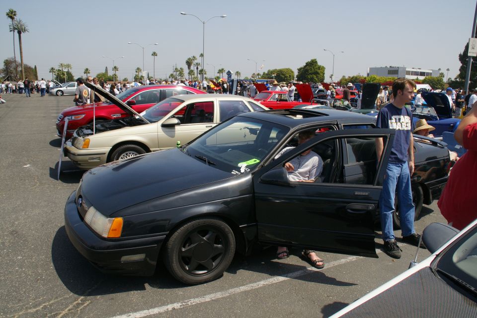 Black sedan with driver's door open, parked in a lot with other cars and people