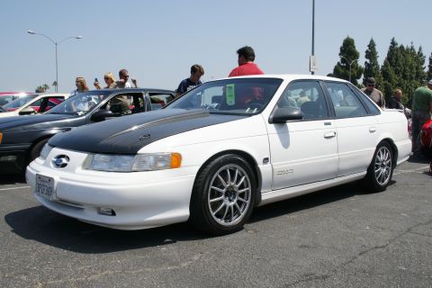 1992 Ford Taurus - A white 1992 Ford Taurus sedan is parked at an outdoor car show, a contrasting black hood and custom wheels.