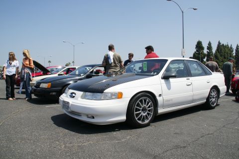1992 Ford Taurus - White Ford Taurus with aftermarket hood and wheels at a car show event.