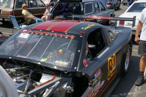 1992 Ford Taurus - A black race car with racing decals and a large rear spoiler is parked at an event, with other vehicles visible in the background.