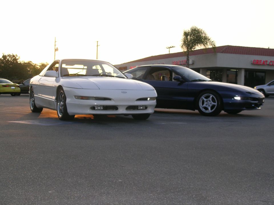 White Ford Probe parked in front of a dark blue Ford Probe, with a building and palm tree in the background